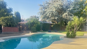 Una piscina con borde curvo, rodeada por una terraza de concreto. Se observan dos sillas reclinables y un pequeño pabellón al fondo.