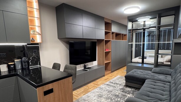 Un salon moderne avec un canapé gris, une table basse et un téléviseur. On y trouve également un coin cuisine doté d’un plan de travail noir et d’armoires en bois.