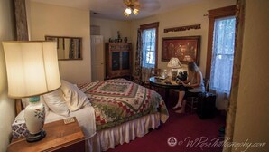 Ein Schlafzimmer mit Bett, Schreibtisch, Stuhl, Lampe, Spiegel und einem Fenster mit Vorhängen.
