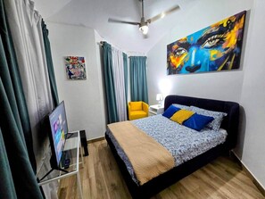 Un dormitorio con cama, televisor, ventilador de techo y un cuadro en la pared.