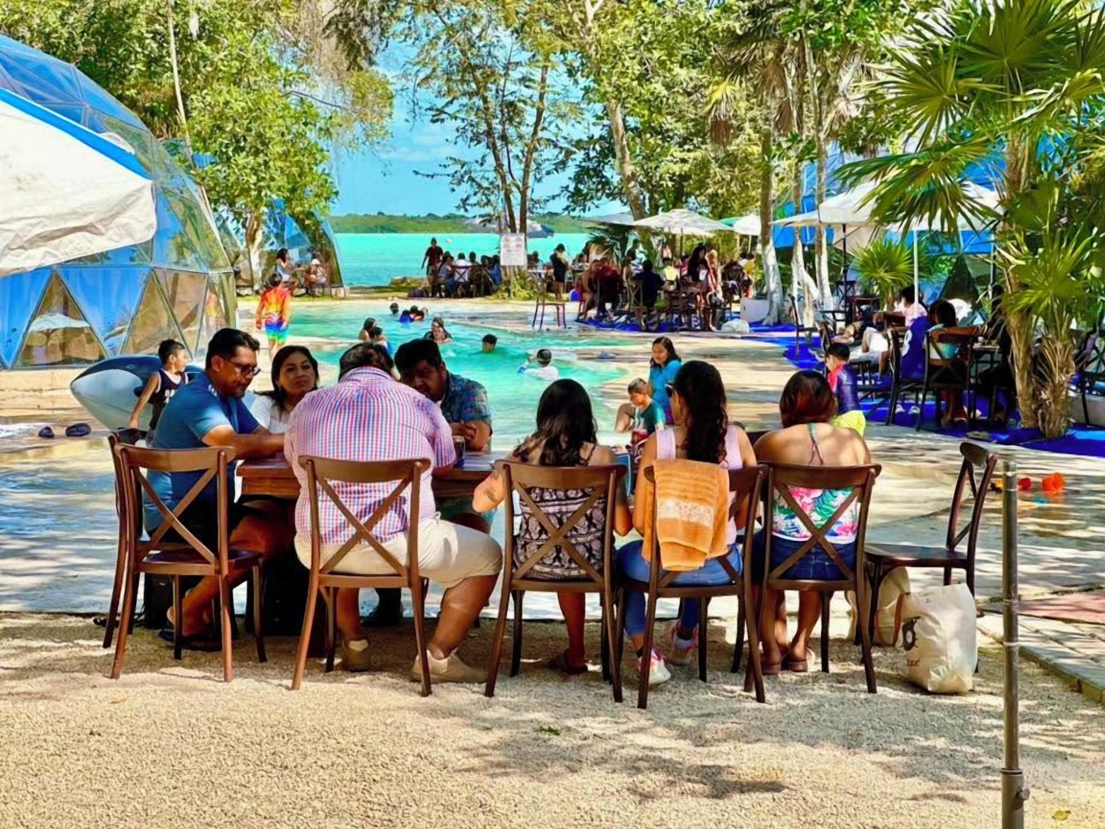 A group of people seated at a table by a poolside, with a view of the pool and the ocean in the background.