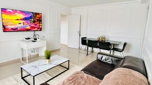 Een moderne woonkamer met een grote flatscreen-tv, een wit bureau, een eethoek met zwarte stoelen en een bruine leren bank.