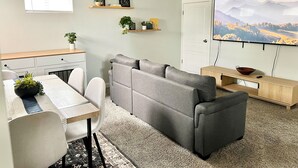 A modern living room with a grey sectional sofa, a dining table with chairs, a small kitchen area, and a large TV mounted on the wall. - Clean Modern Apartment-75” SMART TV & Xbox (Payson)