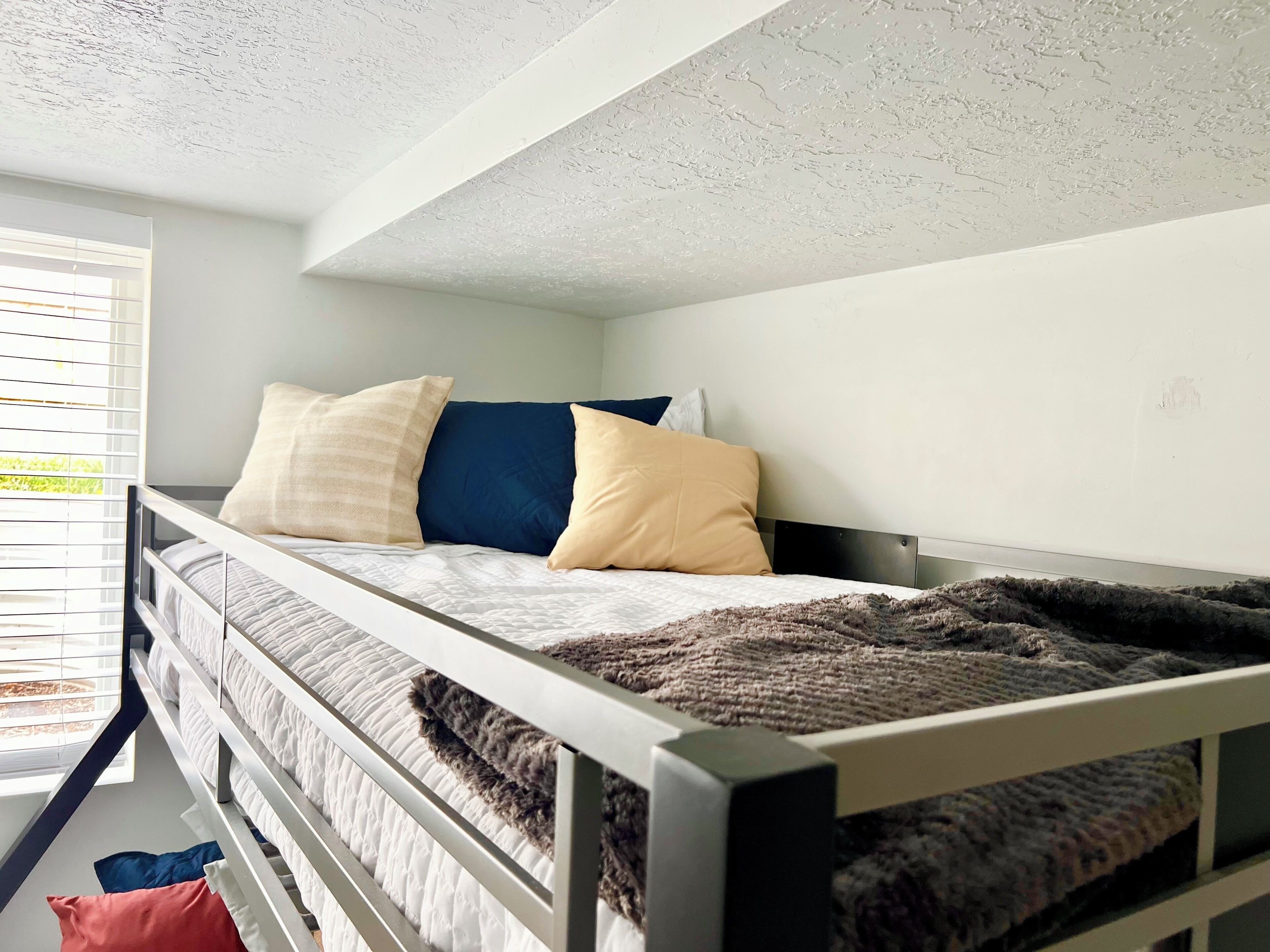 Una cama individual con dos literas, un cabecero blanco, una ventana con persianas y vistas al exterior con vegetación.