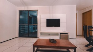 A living room with a flat-screen TV mounted on a white wall, a wooden table with a woven basket, and a sliding glass door leading to a balcony. - Flat Atlantic Ocean - Praia de Iracema (Fortaleza)