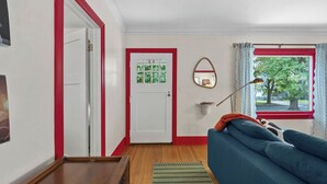 A living room with a blue sofa, a wooden floor, a red-framed door, and a window with a view of trees. - Manito Craftsman with Fire Pit (Spokane)