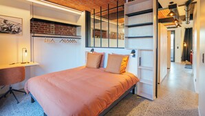 A modern bedroom with a brick ceiling, a bed with orange bedding, a desk with a chair, and a shelving unit. - La Camuche by Hélène Dutrieu (Tournai)