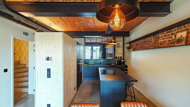 A modern kitchen with a central island, wooden cabinets, and a brick wall. - La Camuche by Hélène Dutrieu (Tournai)