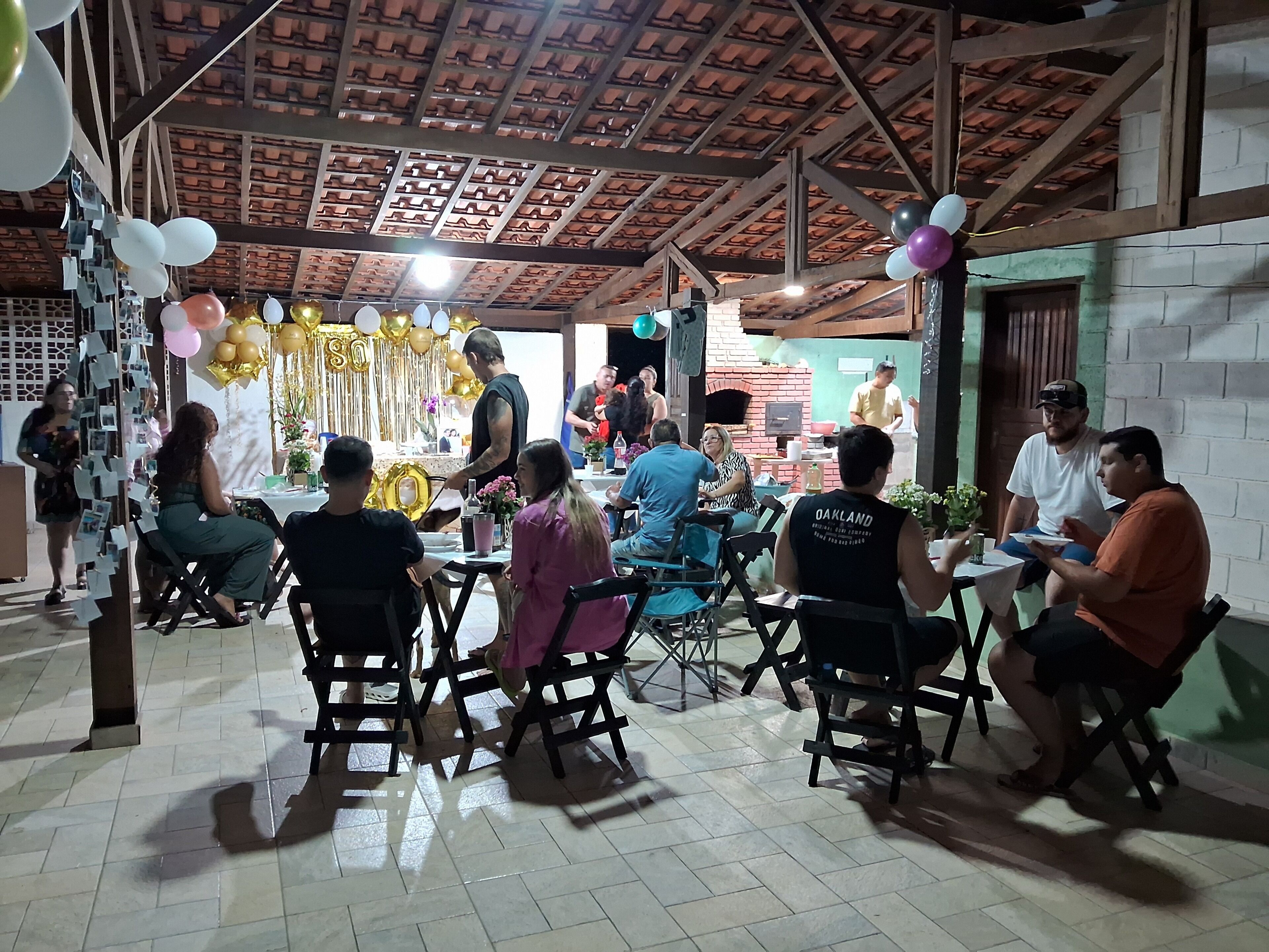 An indoor gathering with people seated at tables, a brick oven in the background, and a ceiling with a wooden pattern.