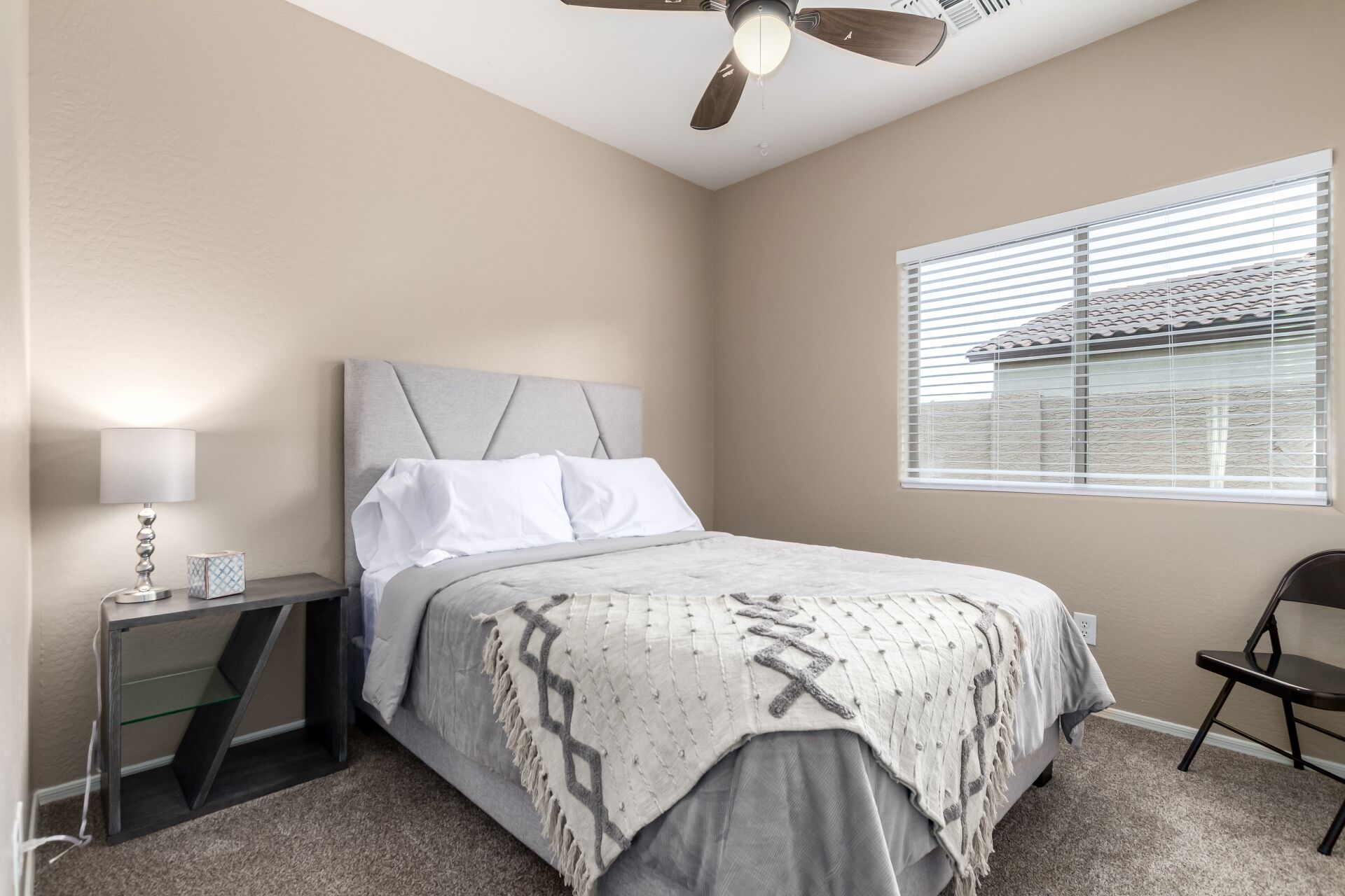 A bedroom with a bed, a nightstand, a lamp, a chair, a window with blinds, and a ceiling fan.