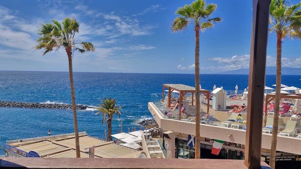 Un balcón con vistas al océano, palmeras y una zona de playa con sombrillas y sillas de descanso.