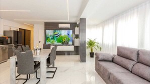 Una sala moderna con un sofá gris, un comedor con mesa y sillas, una cocina con refrigerador y horno, y un televisor mostrando un acuario.