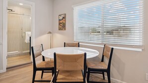 A modern dining area with a round table and four chairs, a large window with blinds, and a shower room with a glass door. - Chic & Cozy Studio | King Bed + Sleeper (Tempe)