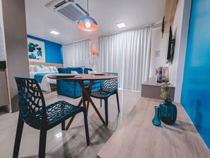 A modern living room with a blue sofa, a dining table with chairs, and a large window with white curtains. - Compacto de luxo - One Milênio Cocô (Fortaleza)