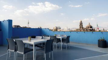A rooftop terrace with a blue wall, a white table, and chairs, overlooking a cityscape with a prominent dome and flag.