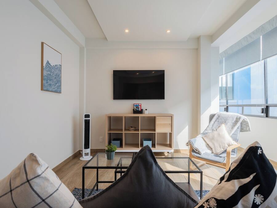 A modern living room with a flat-screen TV mounted on the wall, a wooden shelving unit, and a comfortable seating area with a chair and cushions.