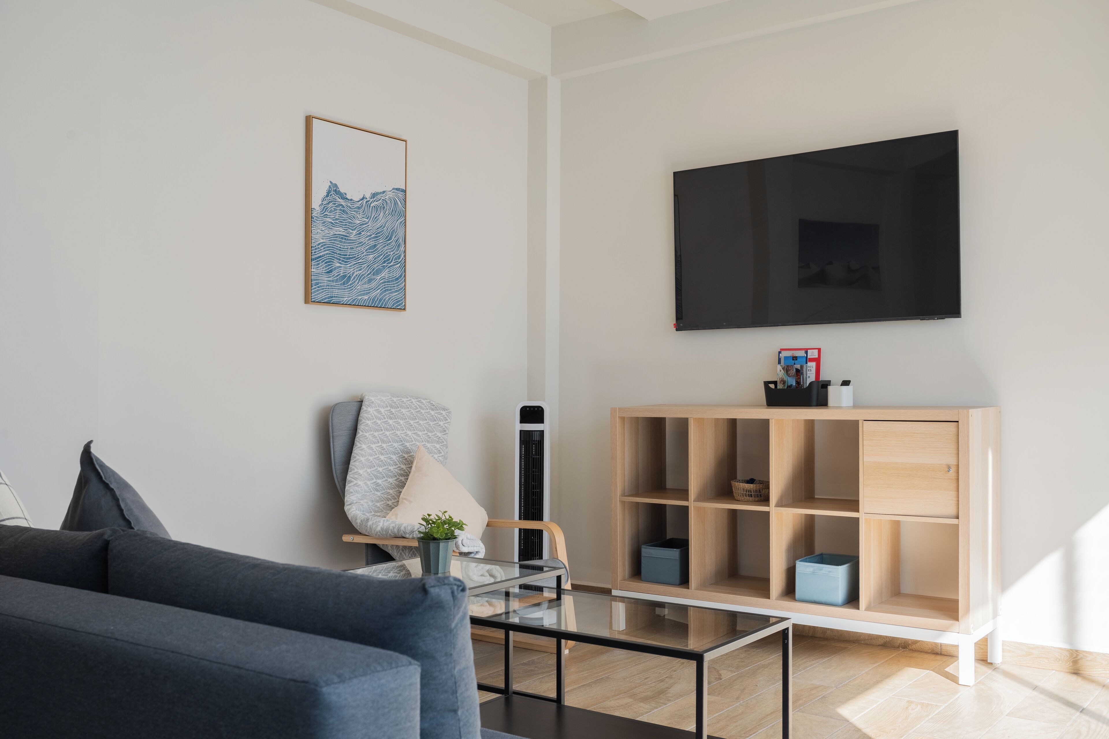 Un salon moderne avec un canapé gris, une table basse en verre, une bibliothèque en bois, un téléviseur fixé au mur et un tableau abstrait encadré.