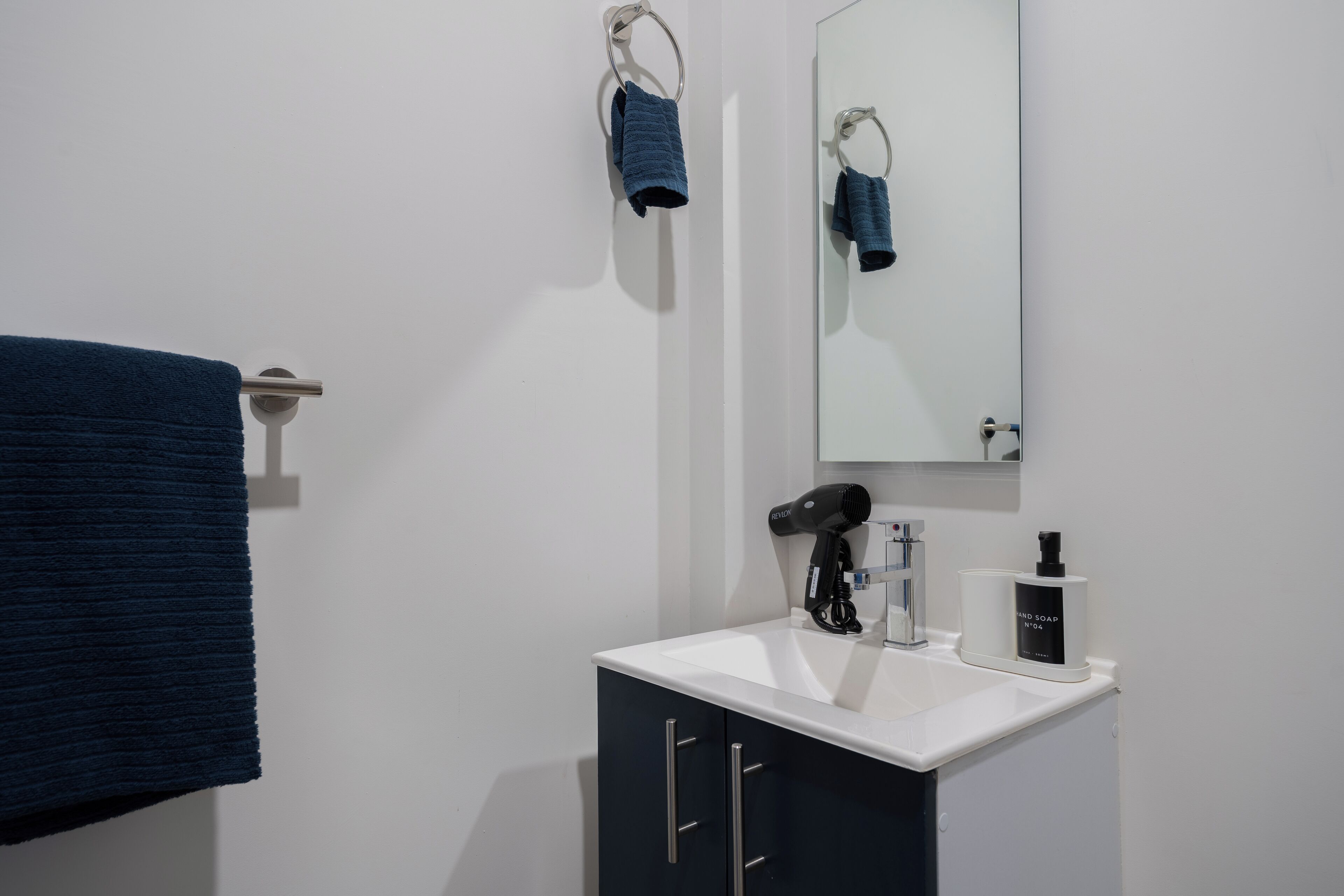 A bathroom with a white sink, dark cabinets, a towel rack, a hairdryer, and a soap dispenser.