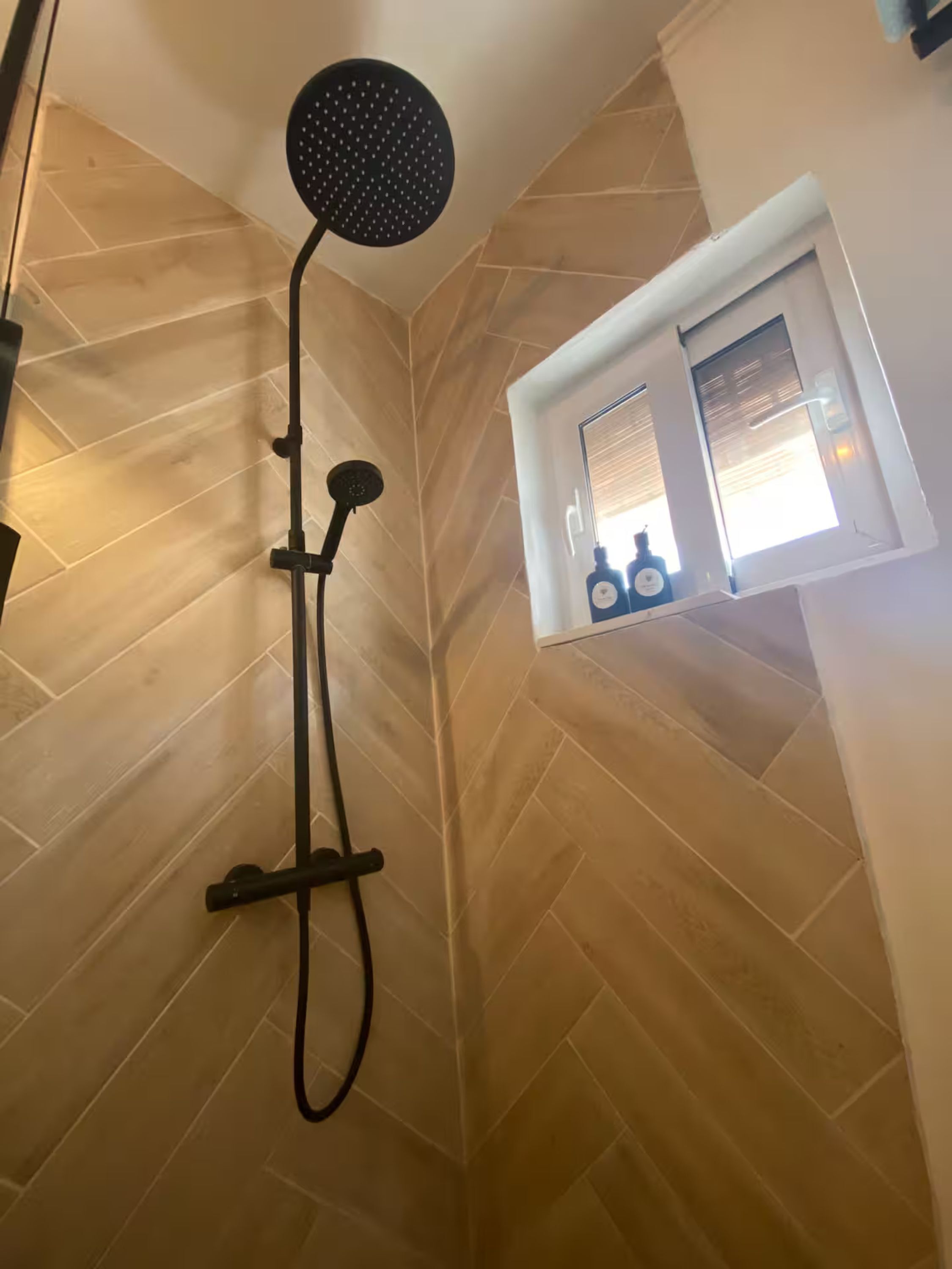 A shower with a black head, a handheld showerhead, and a wall-mounted control. There is a window with a view of bottles and a bottle of shampoo.