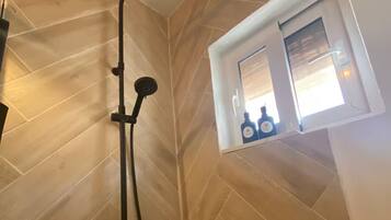 A shower with a black head, a handheld showerhead, and a wall-mounted control. There is a window with a view of bottles and a bottle of shampoo.