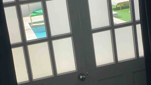 A view through a window with a view of a pool and a grassy area. - Palm Beach Vacaysuites - Garden Suite - Poolside (Noord)