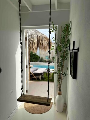 Ein Zimmer mit einer Schaukelstuhlsitzgelegenheit, einer weißen Wand, einer Bambuspflanze und Blick auf einen Pool und Gartenmöbel.