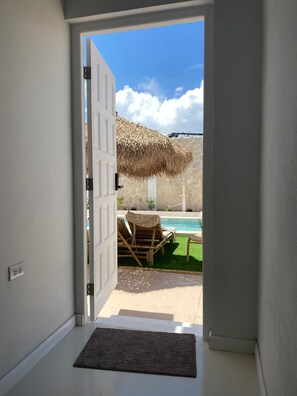 Una entrada que da a una zona de piscina con una sombrilla de paja, sillas de descanso y un muro de piedra.