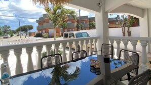 A dining area with a glass table, wicker chairs, and a woven ceiling light. - Palm Beach Vacaysuites - Palm Suite 100m2 (Noord)