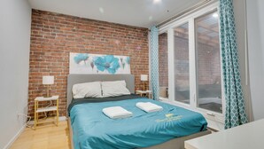 A bedroom with a brick wall, a bed with a blue bedspread, a window with curtains, and a small bedside table with a lamp.