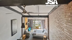 Une chambre d’hôtel moderne avec un lit, une petite table et un canapé. Il y a une fenêtre avec des rideaux, un ventilateur de plafond et un panneau sur le mur.