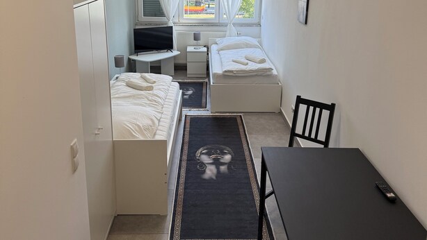 A compact room with a bed, a desk, a chair, and a small table. There is a window with curtains, a radiator, and a painting on the wall. - City apartment no. 1 with comfort (Pforzheim)