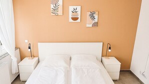 A double bed with white bedding in a room with an orange wall, two bedside tables with lamps, and three wall-mounted artworks. - City apartment no. 1 with comfort (Pforzheim)