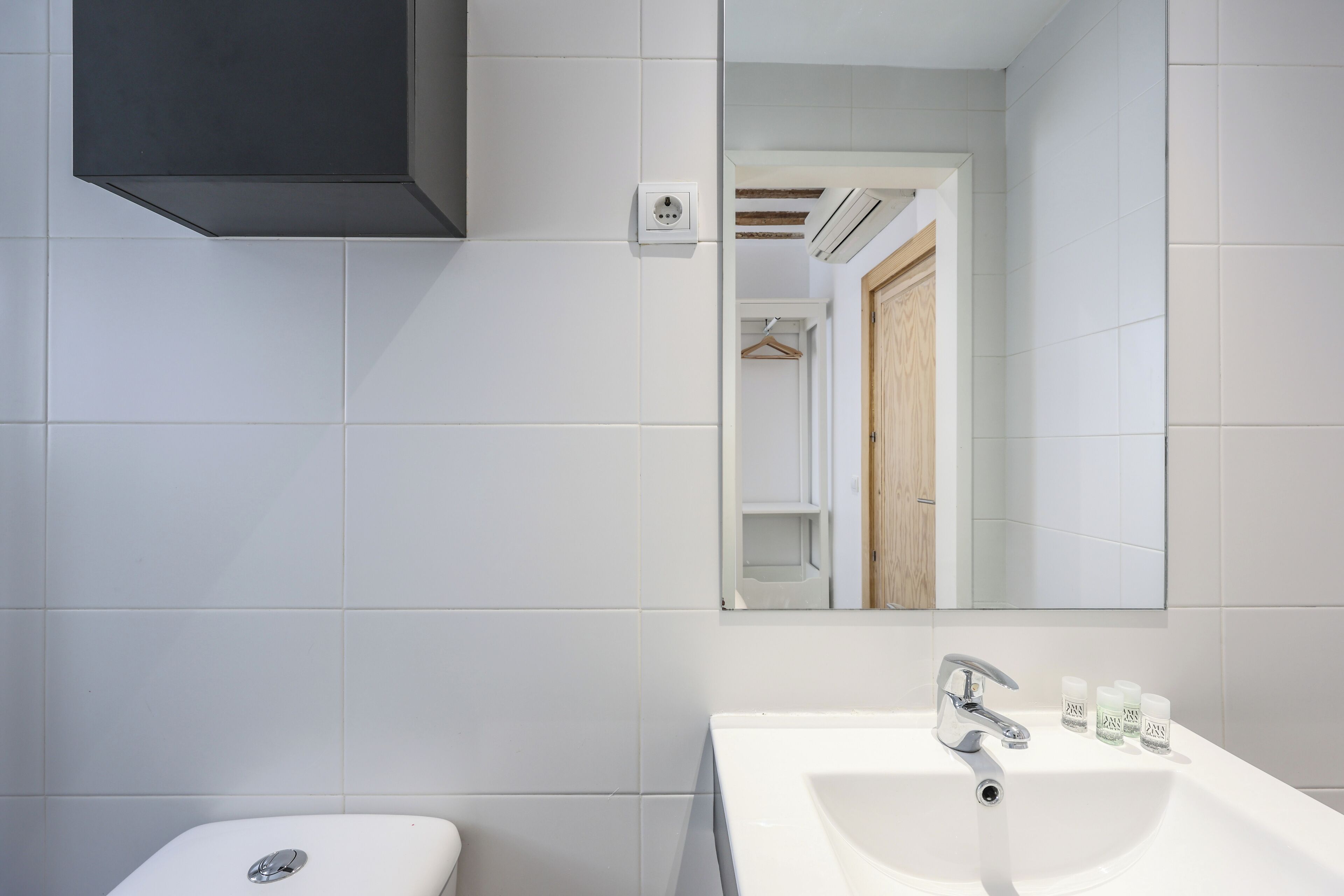 A modern bathroom with white tiles, a white sink, a mirror, and a black cabinet.