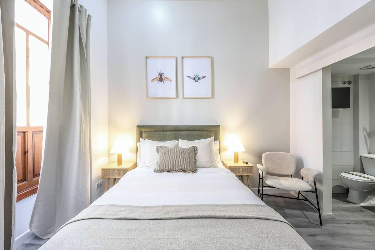 Un lit bien fait, avec deux oreillers, une table de chevet sur laquelle se trouve une lampe, une chaise et deux tableaux encadrés accrochés au mur.