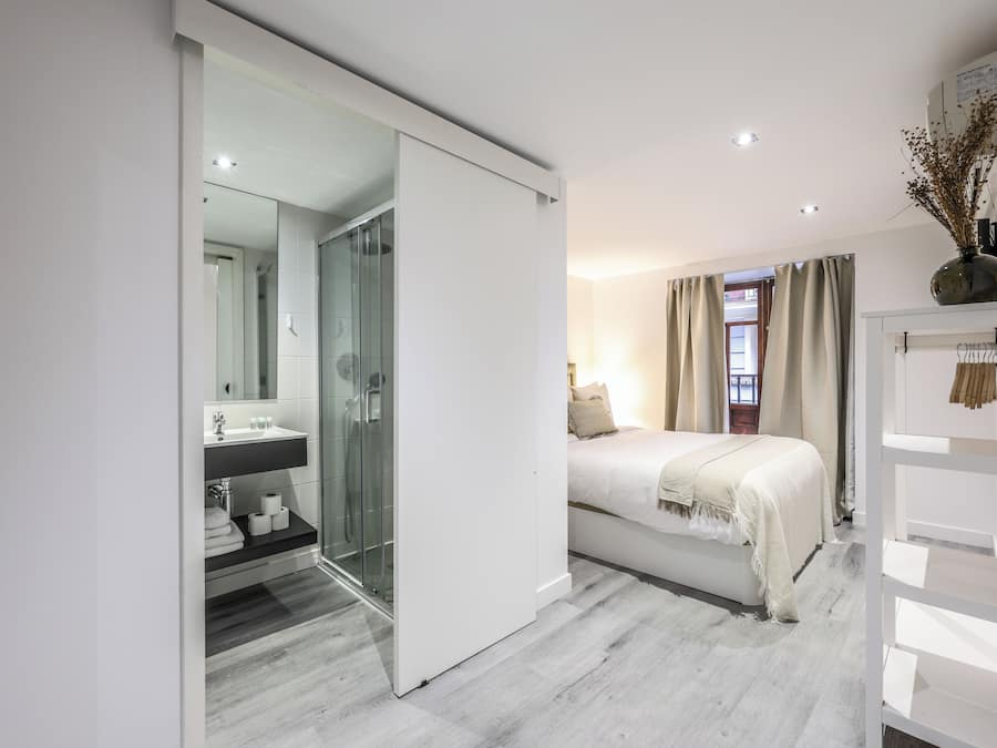 A modern bedroom with a glass shower, a white bed, and a bedside table.