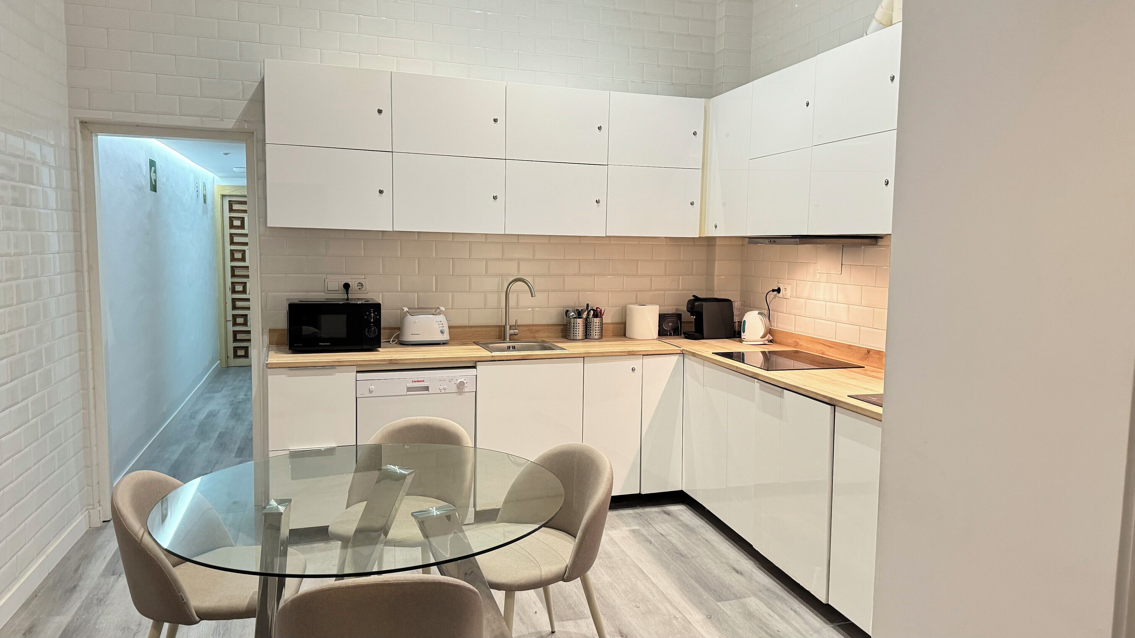 Een kleine keuken met witte kastjes, een magnetron, een broodrooster en een spoelbak. Er staat een glazen tafel met stoelen en een deur naar een andere kamer.