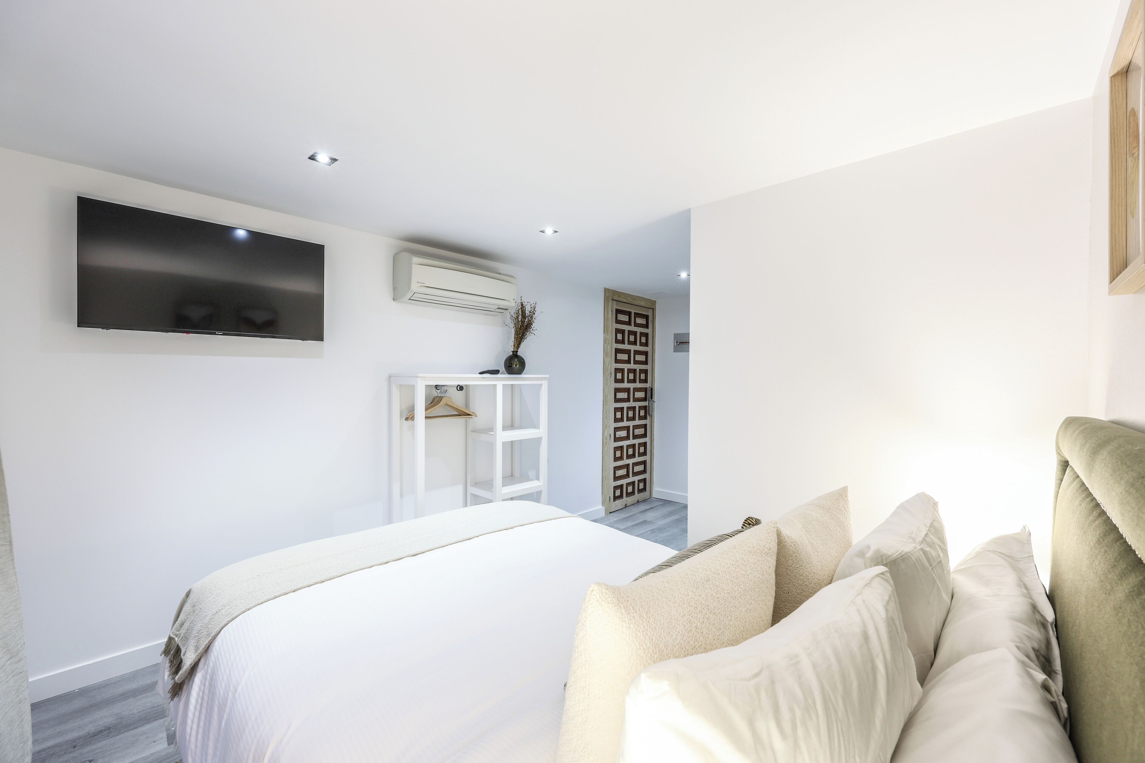 A bedroom with a large bed, a flat-screen TV mounted on the wall, a white bedside table, and a wardrobe.
