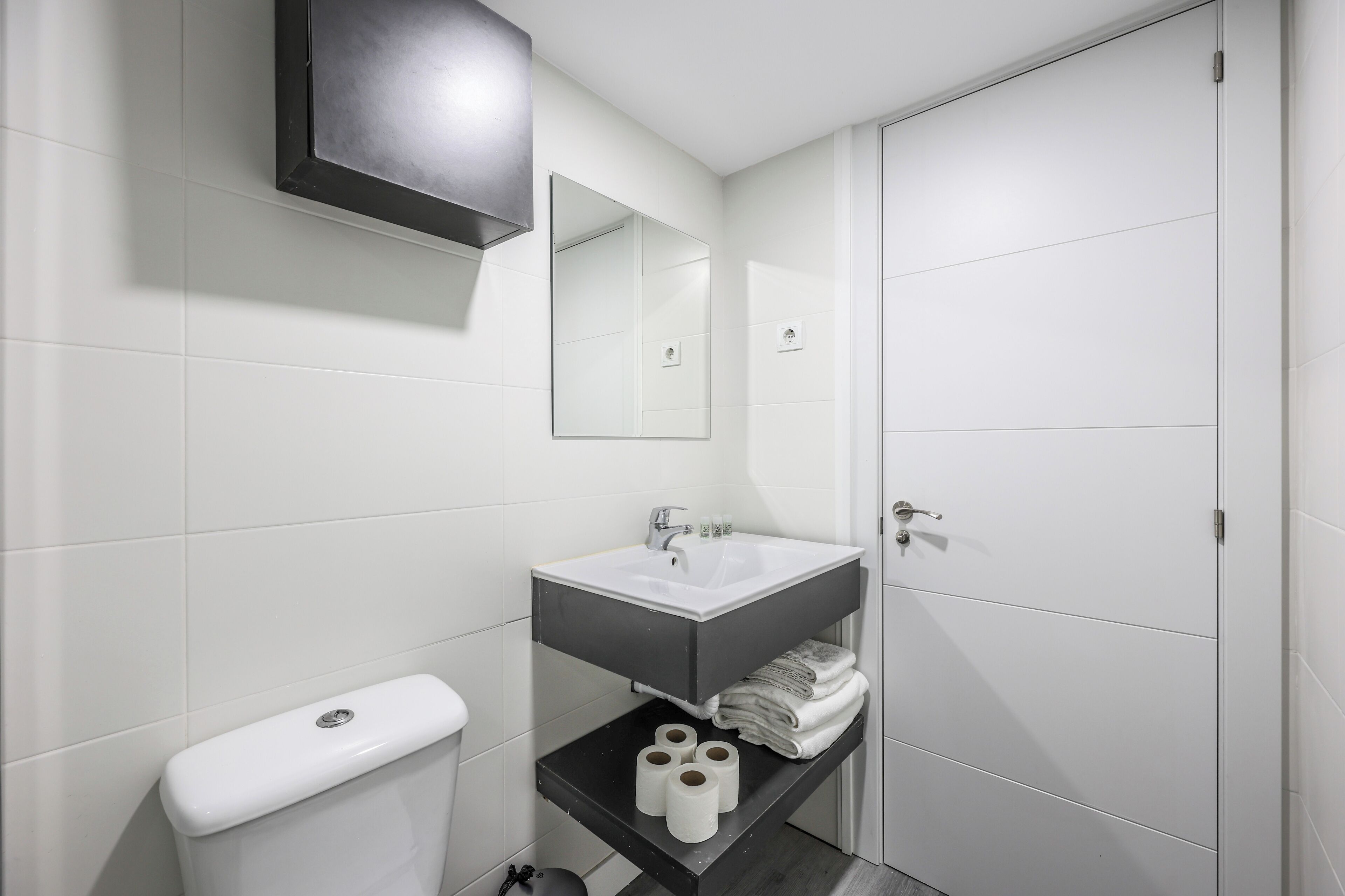 Un baño moderno con inodoro blanco, lavamanos con espejo y estante con toallas y papel higiénico.