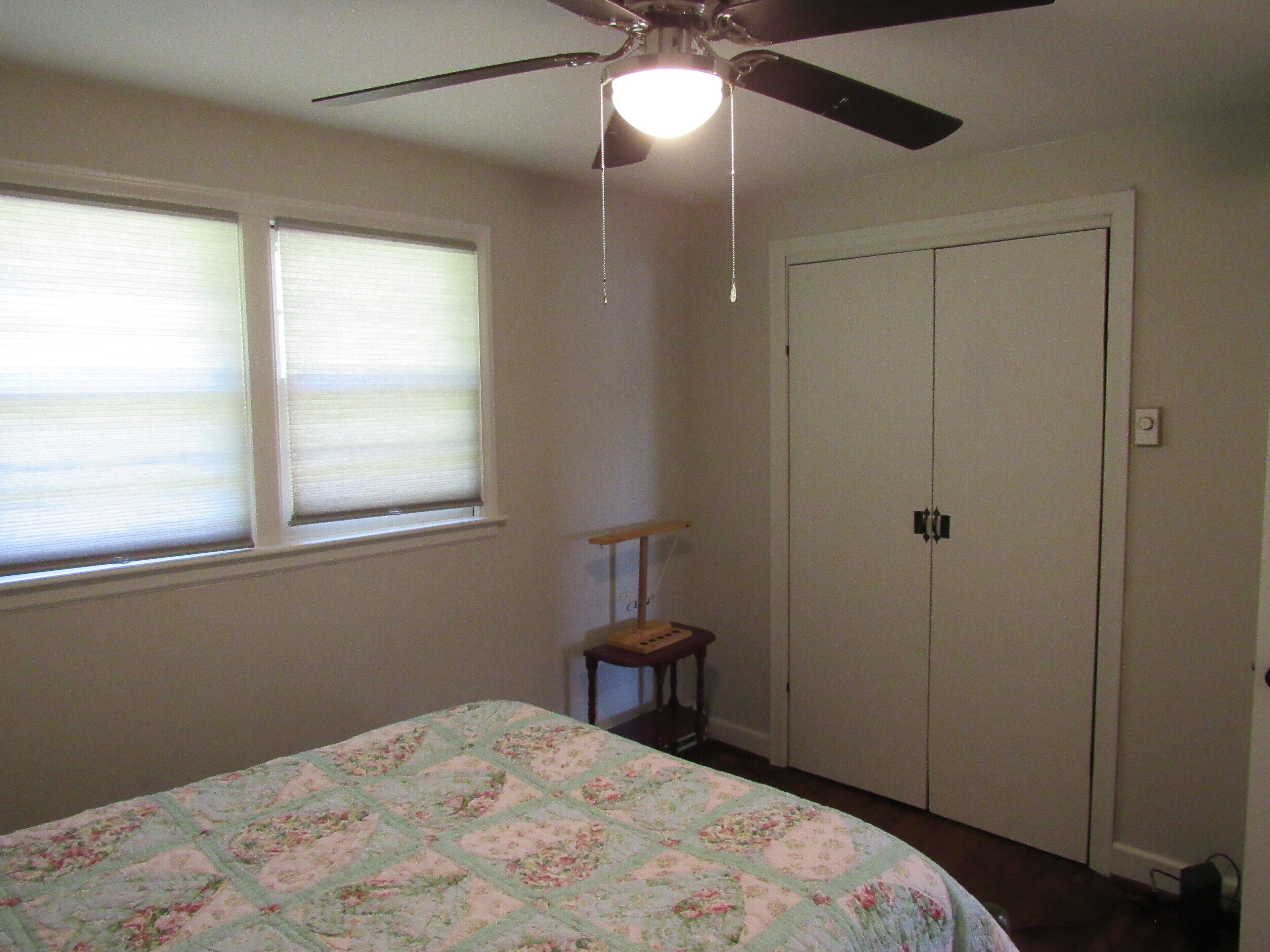 Un dormitorio con cama, ventilador de techo, ventana con persianas, puerta y una silla.