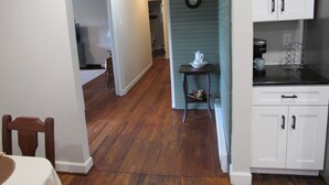 Uma cozinha com piso de madeira, armários brancos e uma pequena mesa com chaleira e xícara.