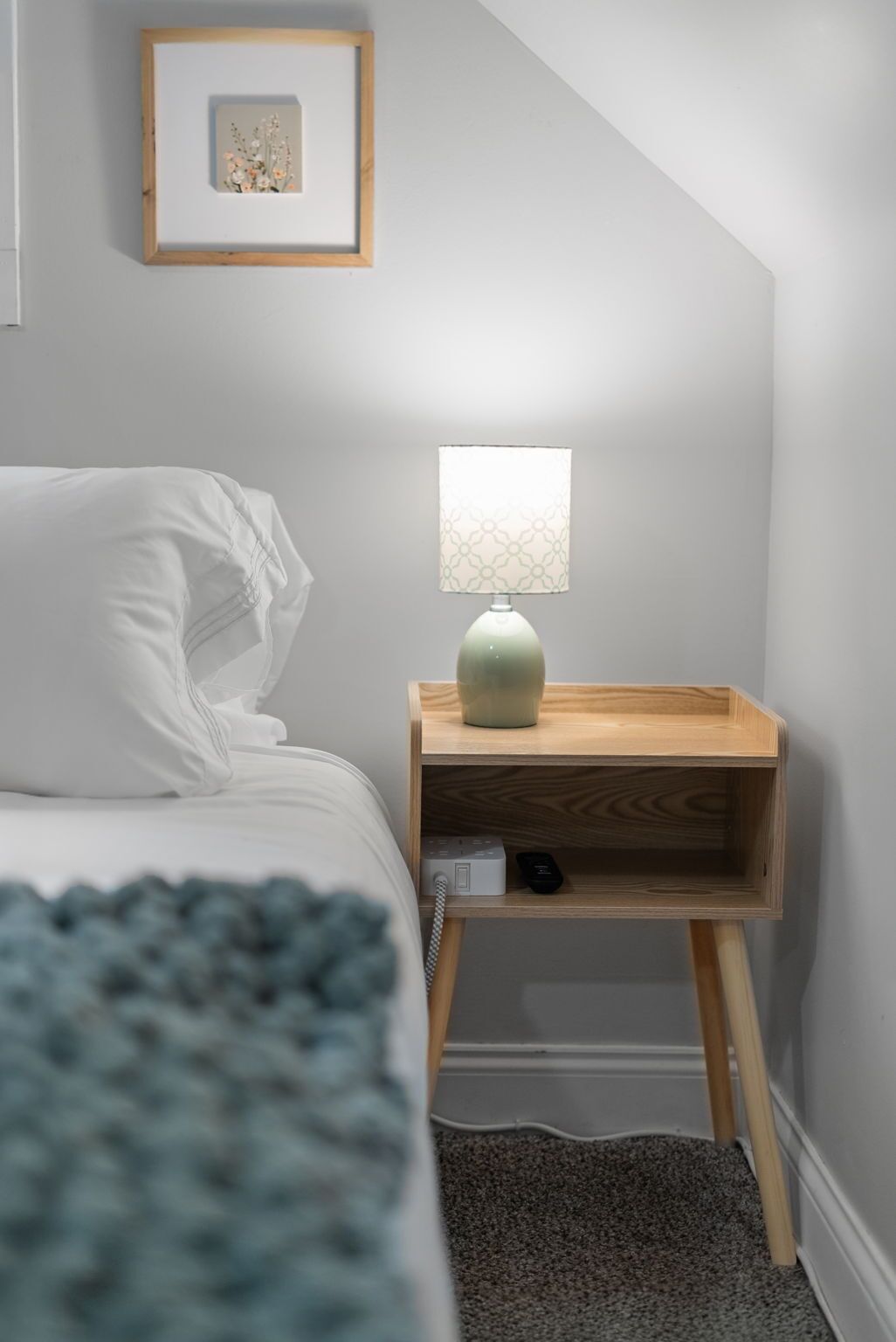 A bedroom with a bed, a bedside table with a lamp, a framed picture on the wall, and a small wooden nightstand.