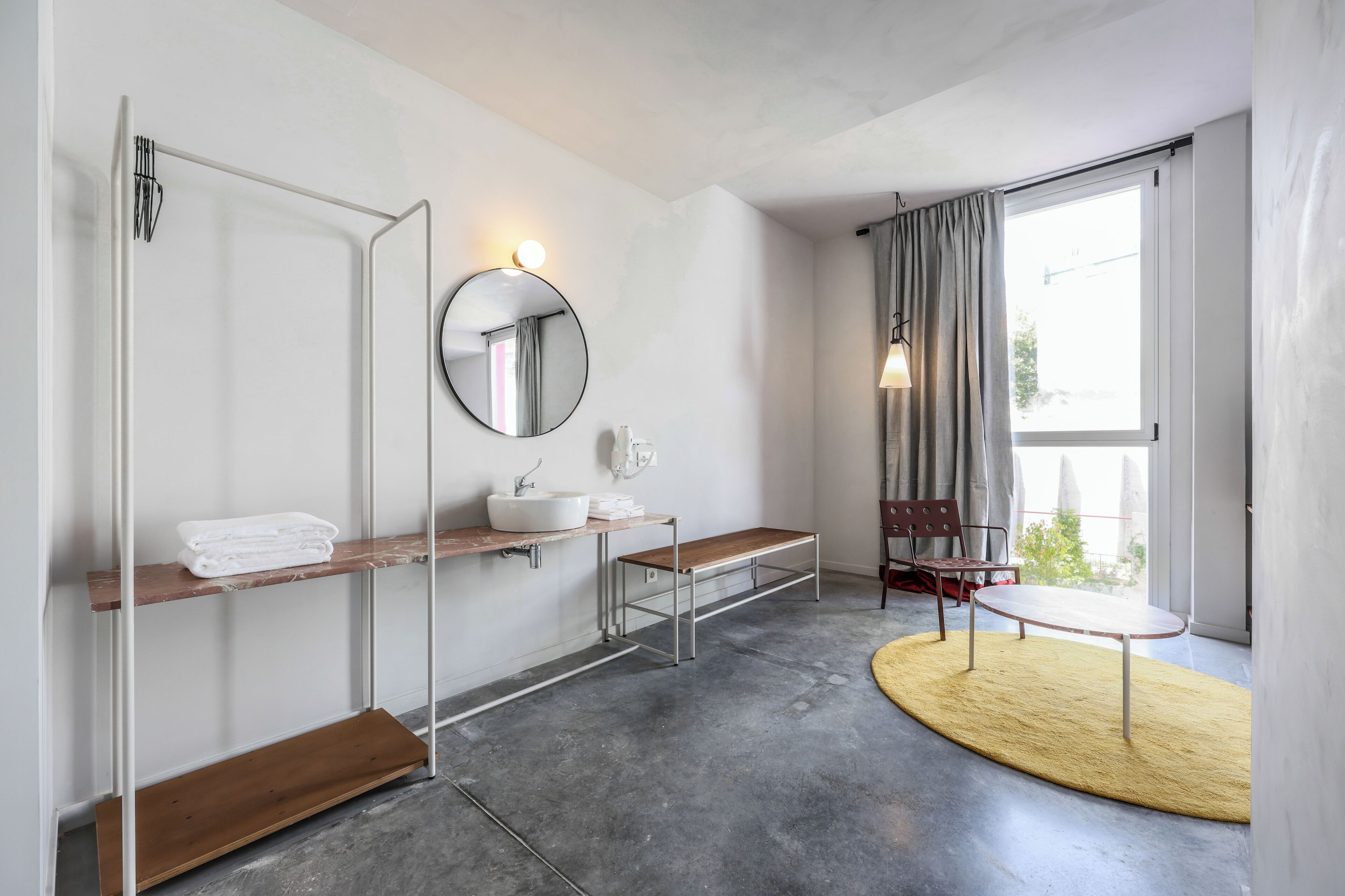Un baño moderno con un espejo redondo, un lavamanos blanco y un banco. Hay una ventana con cortinas y vistas al exterior.