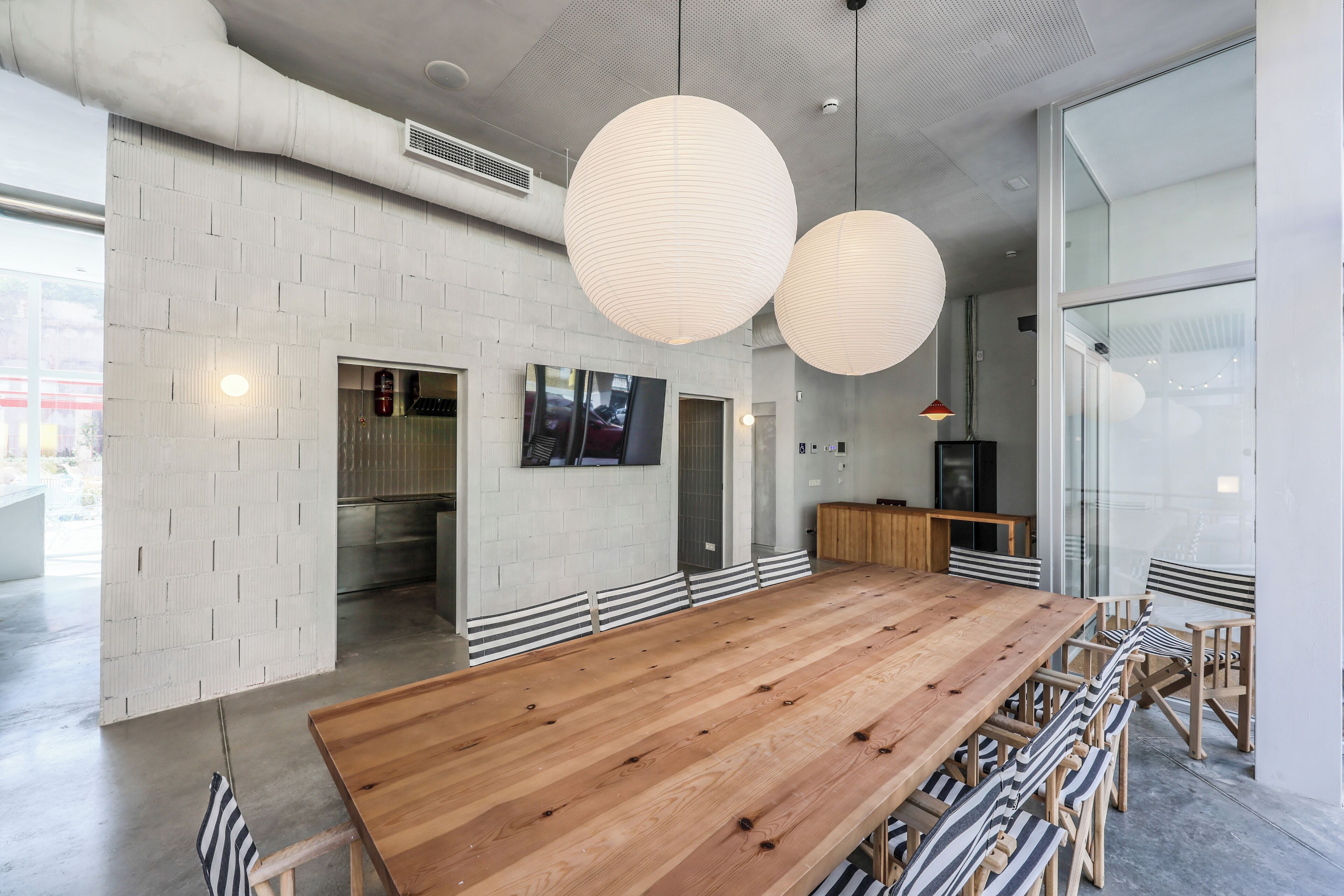 A spacious dining area with a long wooden table and striped chairs, large hanging paper lanterns, and a modern kitchen in the background.