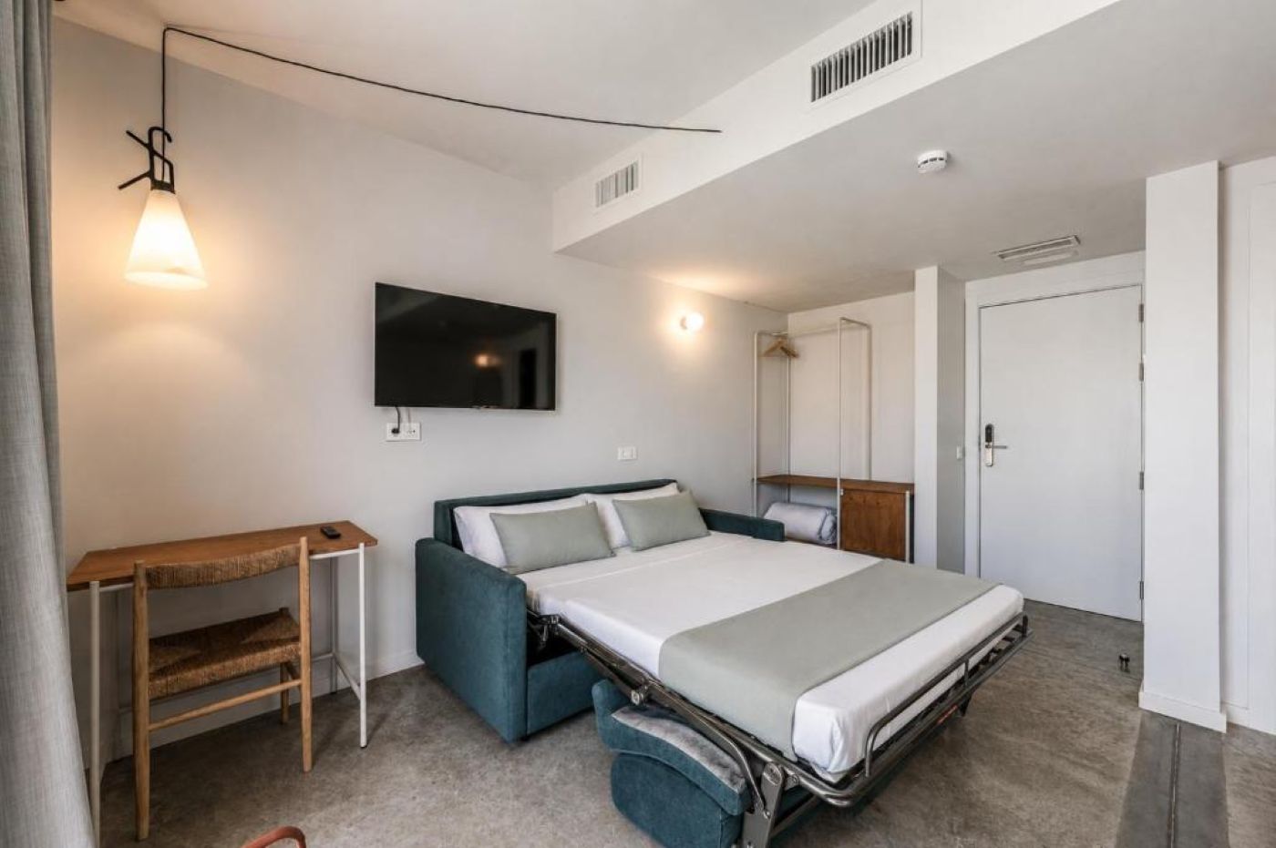 A modern hotel room with a bed, a TV mounted on the wall, a desk, and a wardrobe.