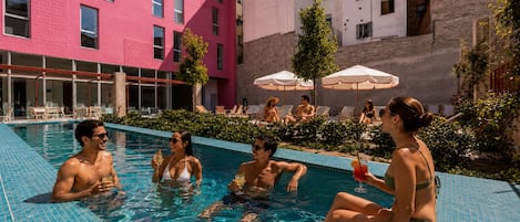 A swimming pool with people relaxing and sunbathing, surrounded by buildings and outdoor seating.