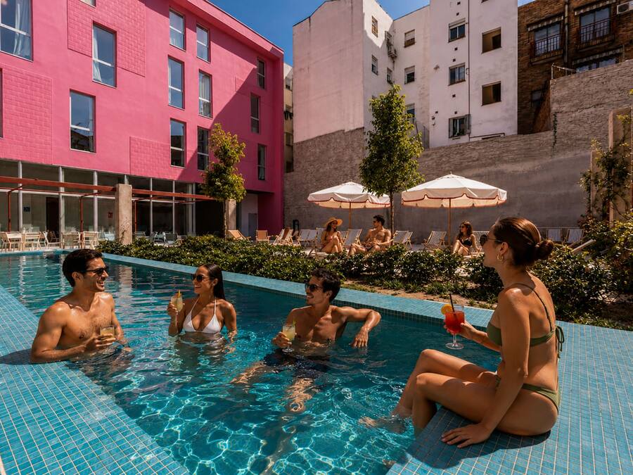 A swimming pool with people relaxing and sunbathing, surrounded by buildings and outdoor seating.