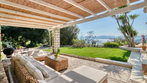 A covered outdoor seating area with wicker furniture and a view of a grassy landscape and water body.
