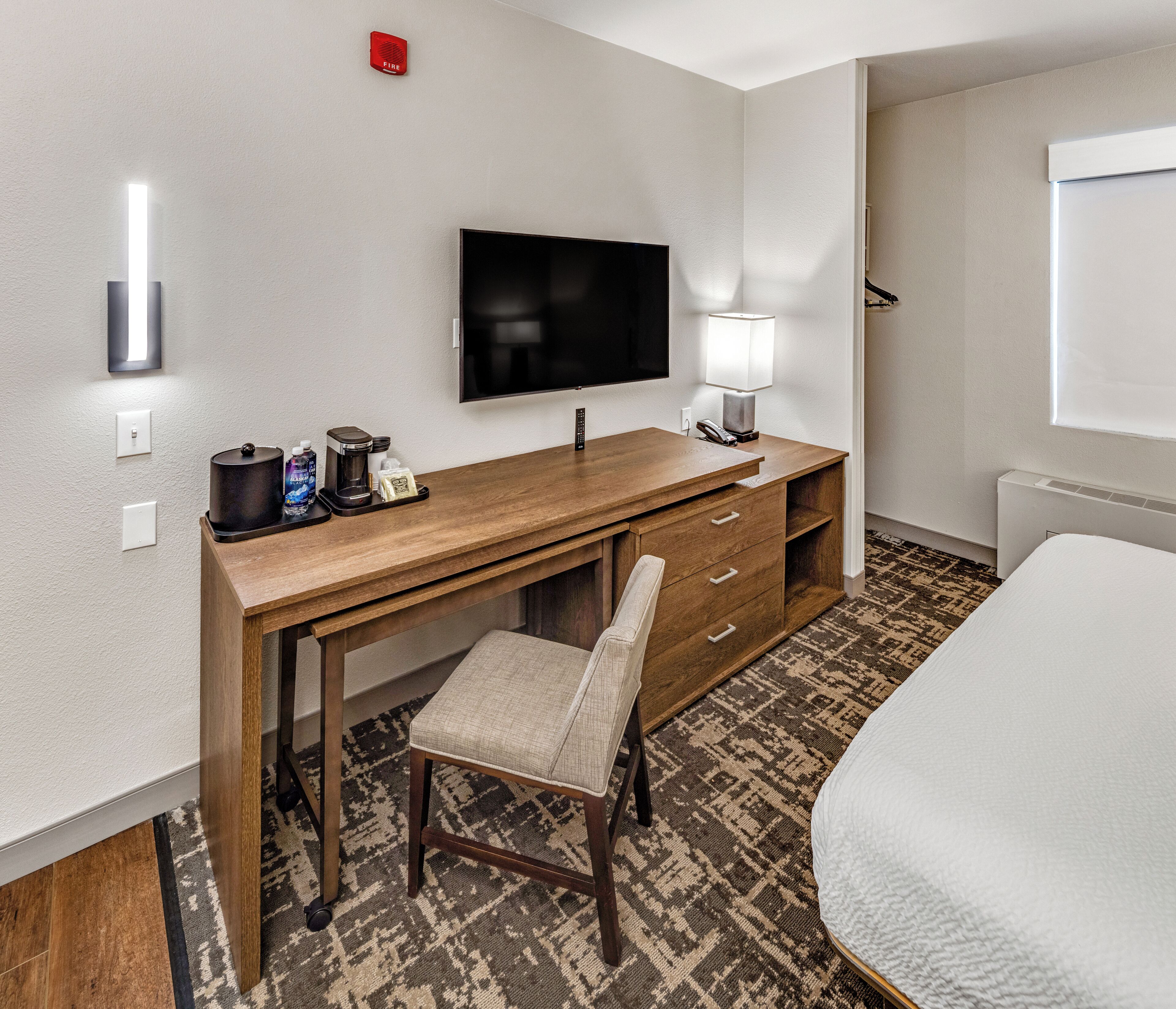 A hotel room with a wooden desk, a television, a chair, a bed, a lamp, and a fire alarm.