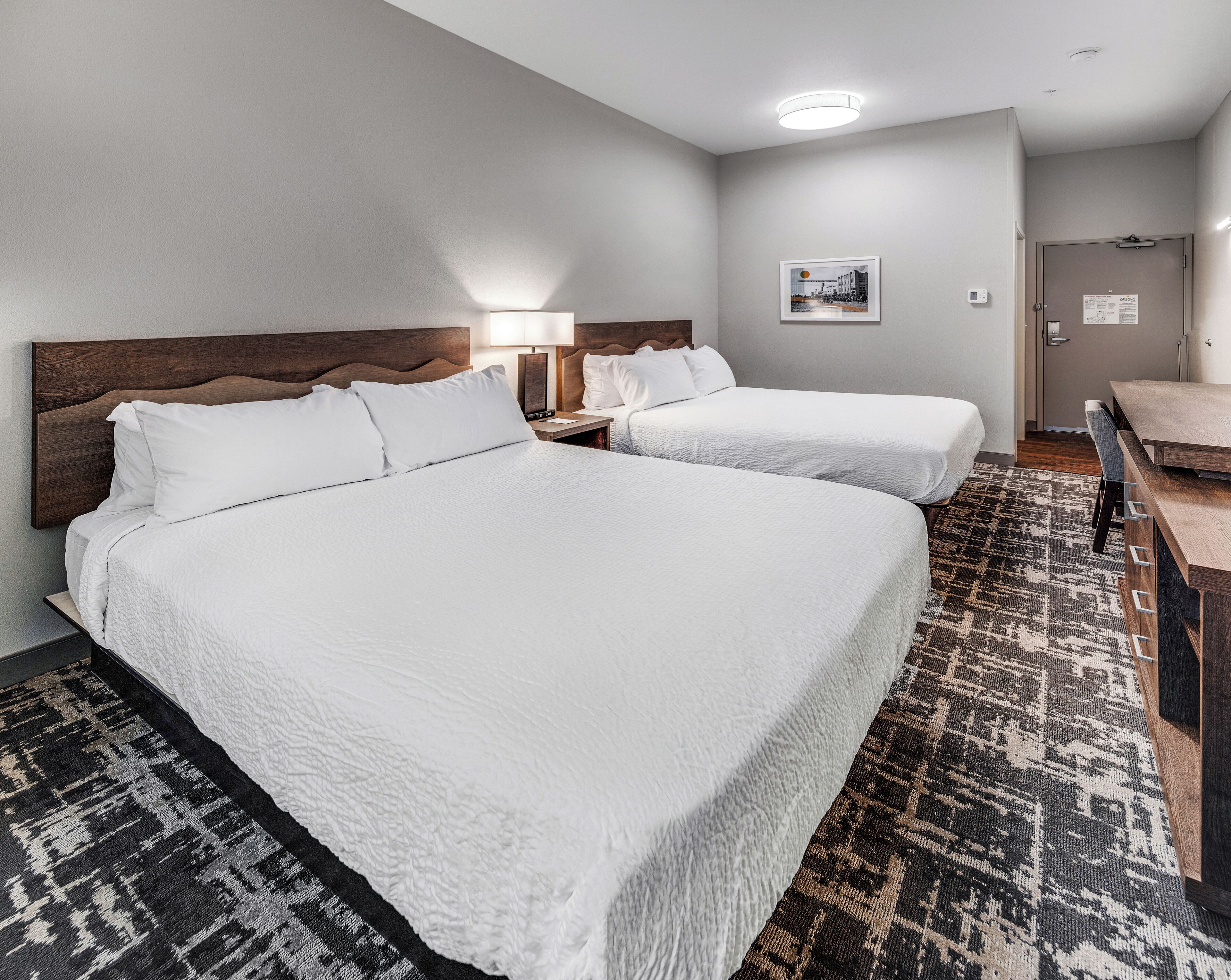 Quarto de hotel com duas camas, uma escrivaninha, uma cadeira, um abajur e um quadro na parede.