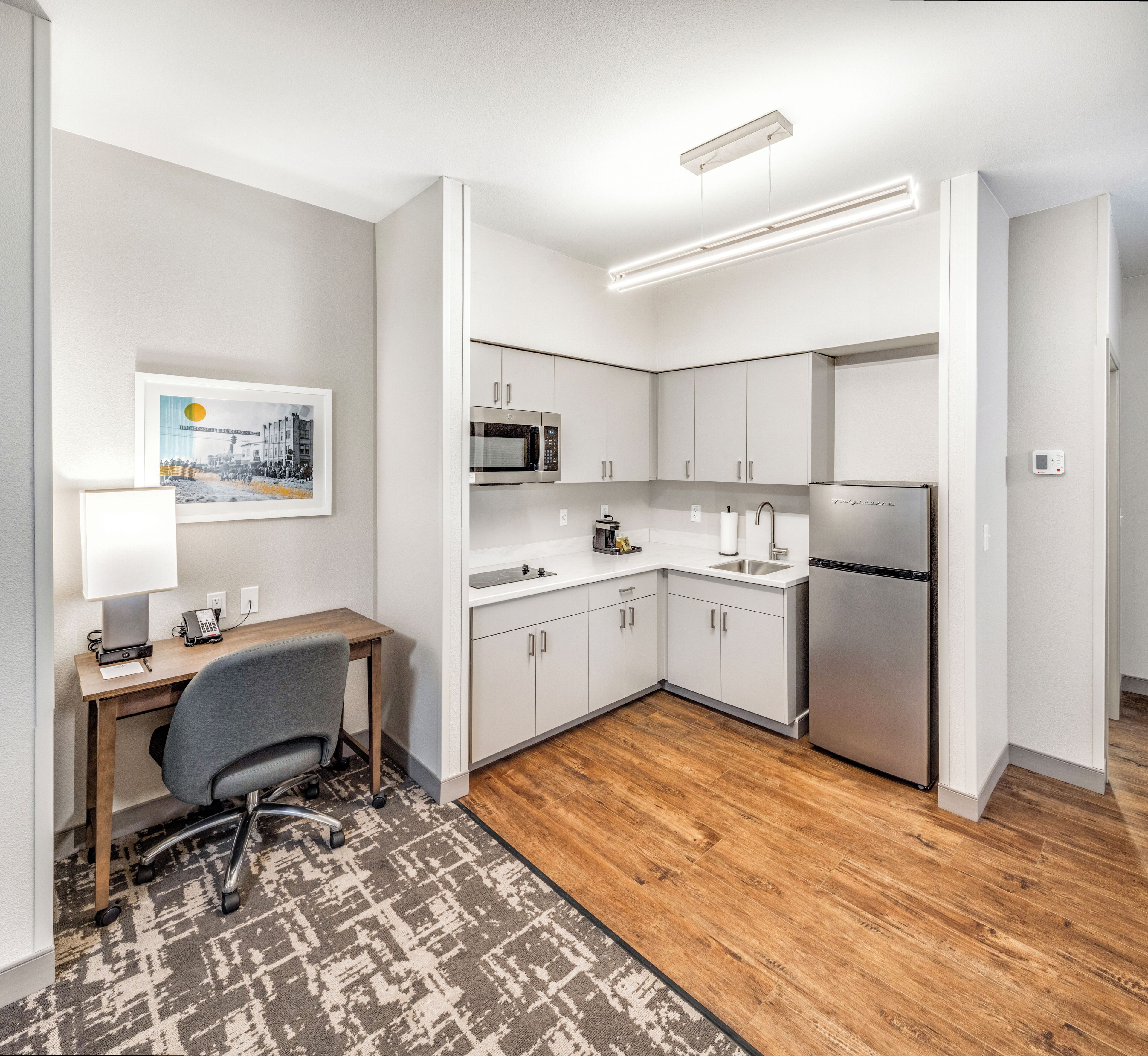 A compact kitchen with white cabinets, a microwave, a refrigerator, and a small dining area with a desk and chair.